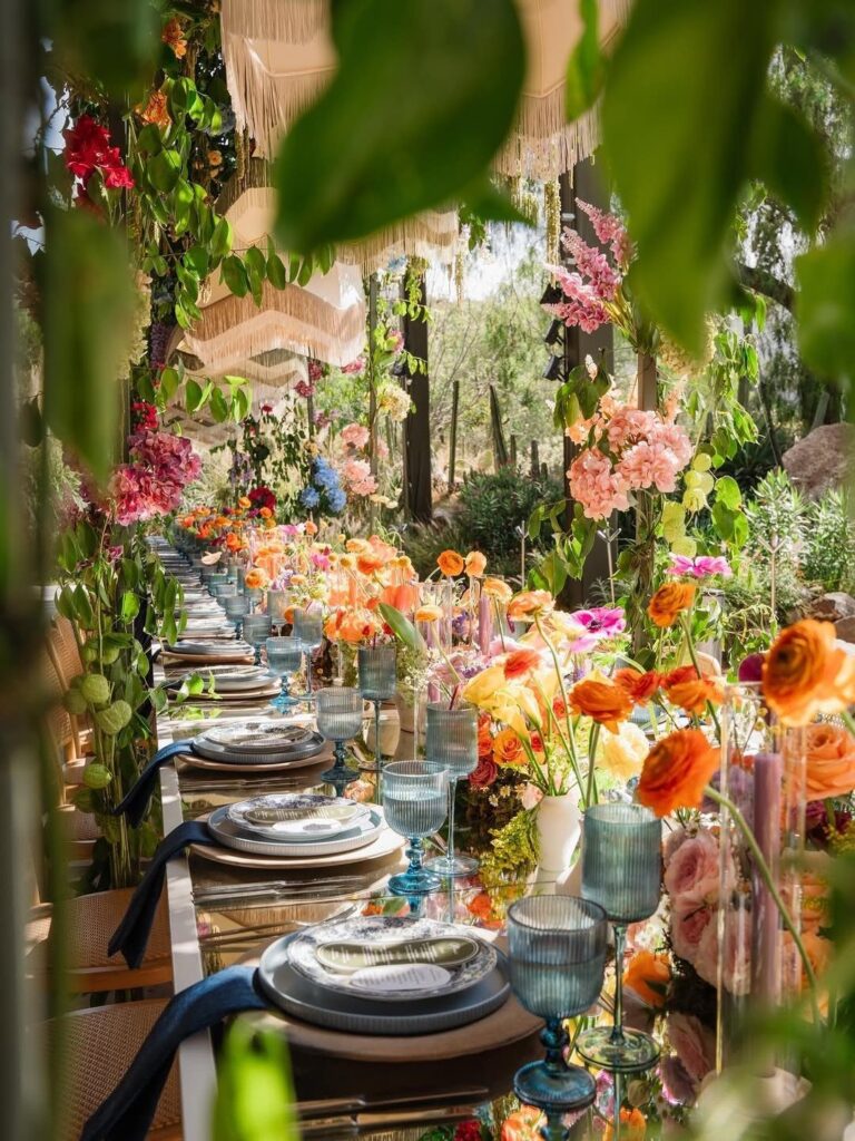 A long outdoor dining table decorated with colorful flowers, elegant glassware, and layered plates.
