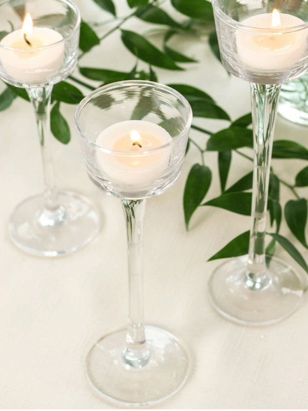 Three lit tea light candles in tall, clear glass stemmed holders among green leaves.