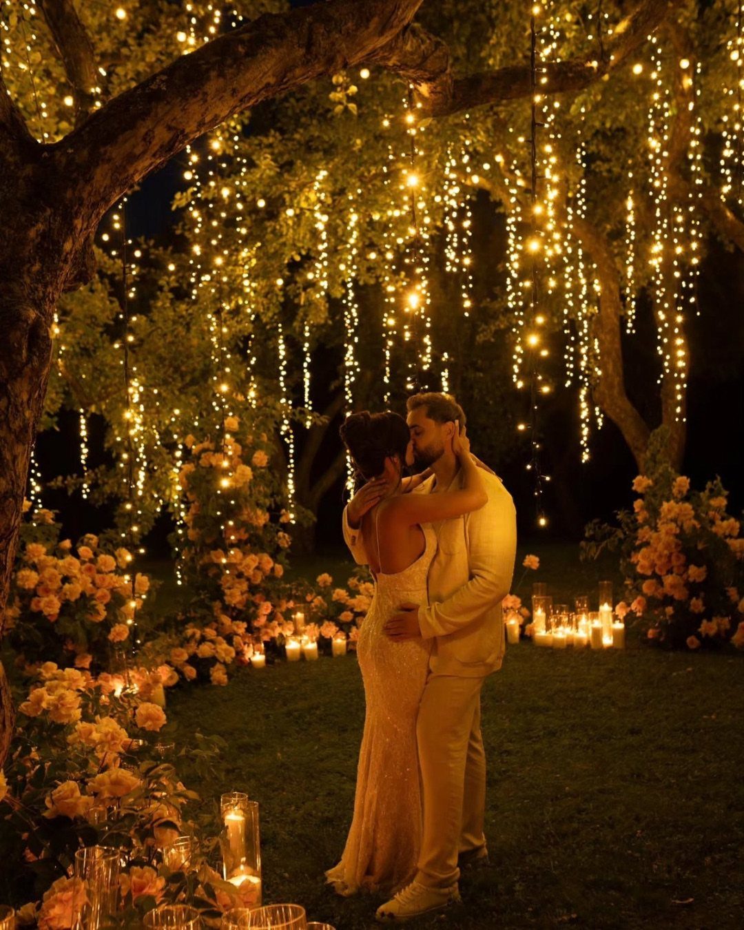 A bride and groom embracing under a tree decorated with cascading fairy lights and flowers.