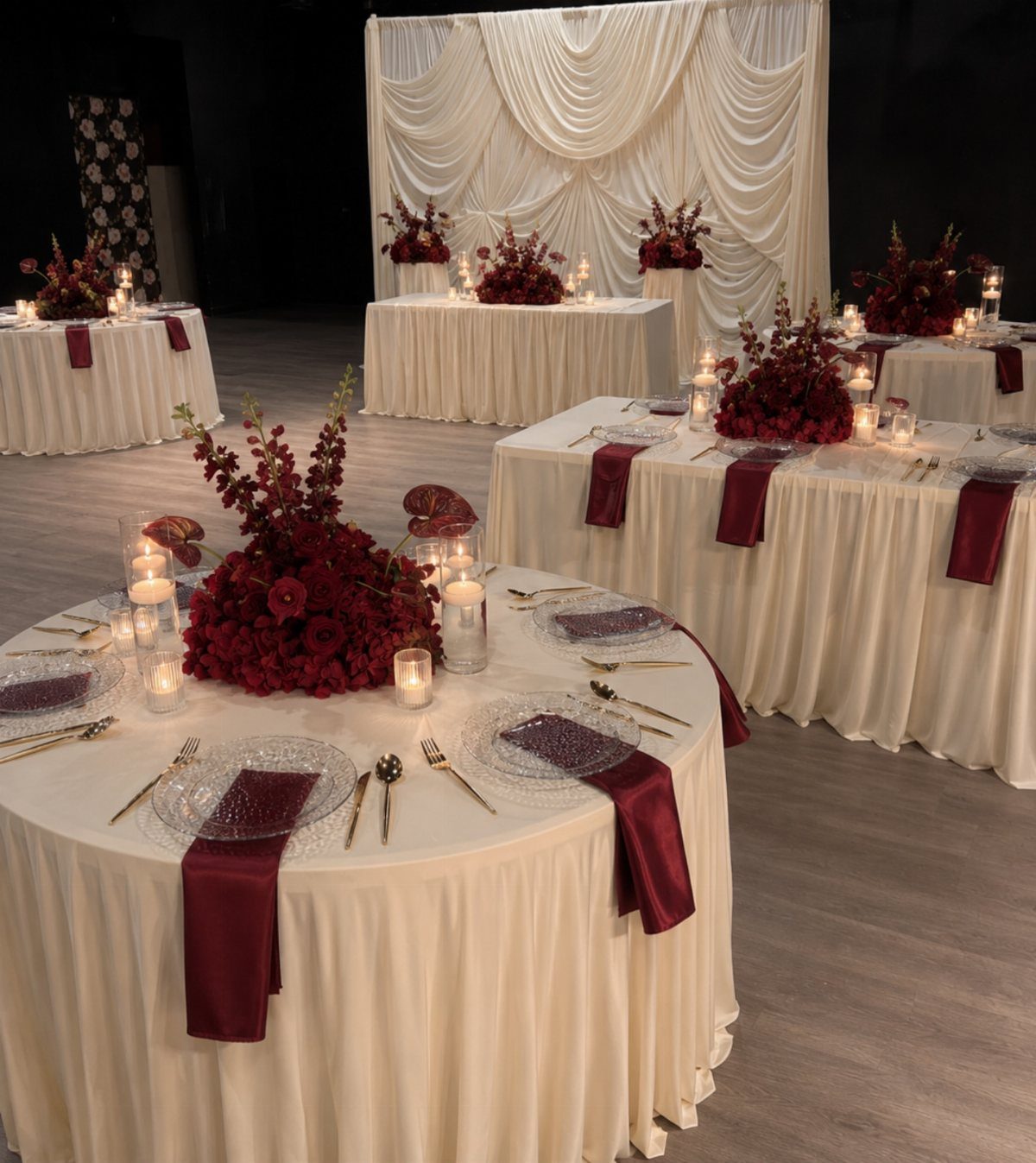 Wedding reception tables featuring cream linens, red floral centerpieces, and burgundy napkins in a dimly lit room.