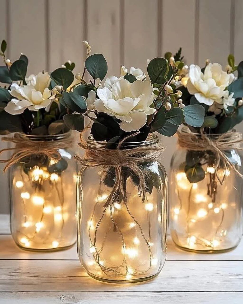 Three mason jars containing white flowers, greenery, and glowing fairy lights on a white surface.