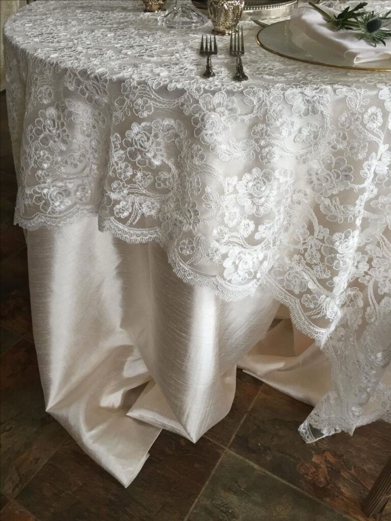 White lace tablecloth with floral patterns draped over a cream satin tablecloth on a set table.