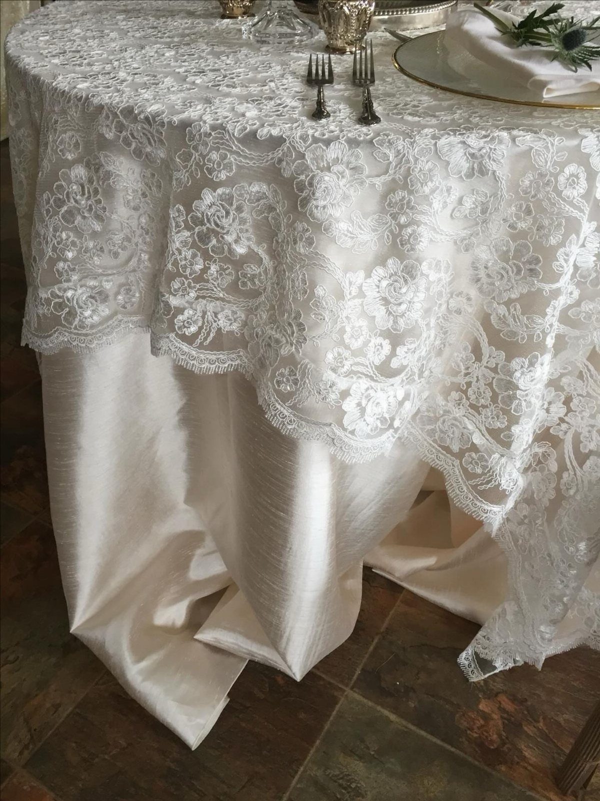 White lace tablecloth with floral patterns draped over a cream satin tablecloth on a set table.