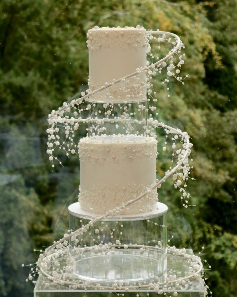 A two-tier white pearl-decorated wedding cake on clear acrylic stands with a beaded vine spiral.