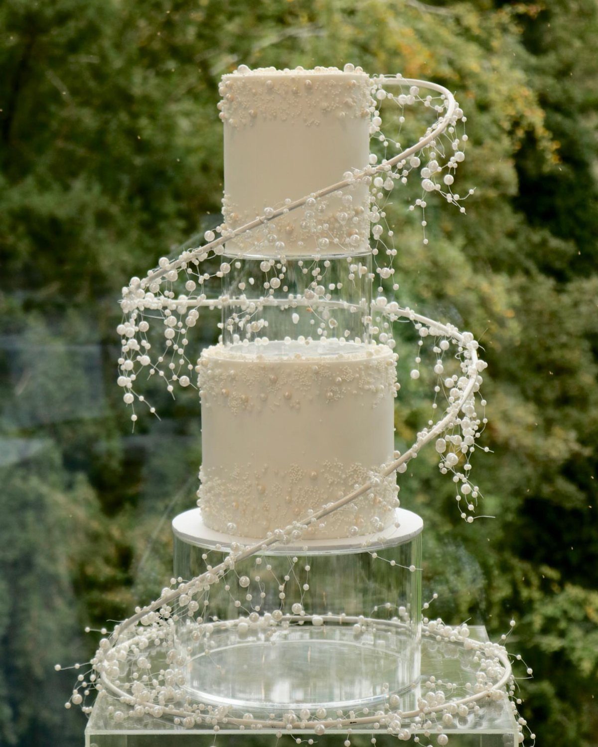 A two-tier white pearl-decorated wedding cake on clear acrylic stands with a beaded vine spiral.