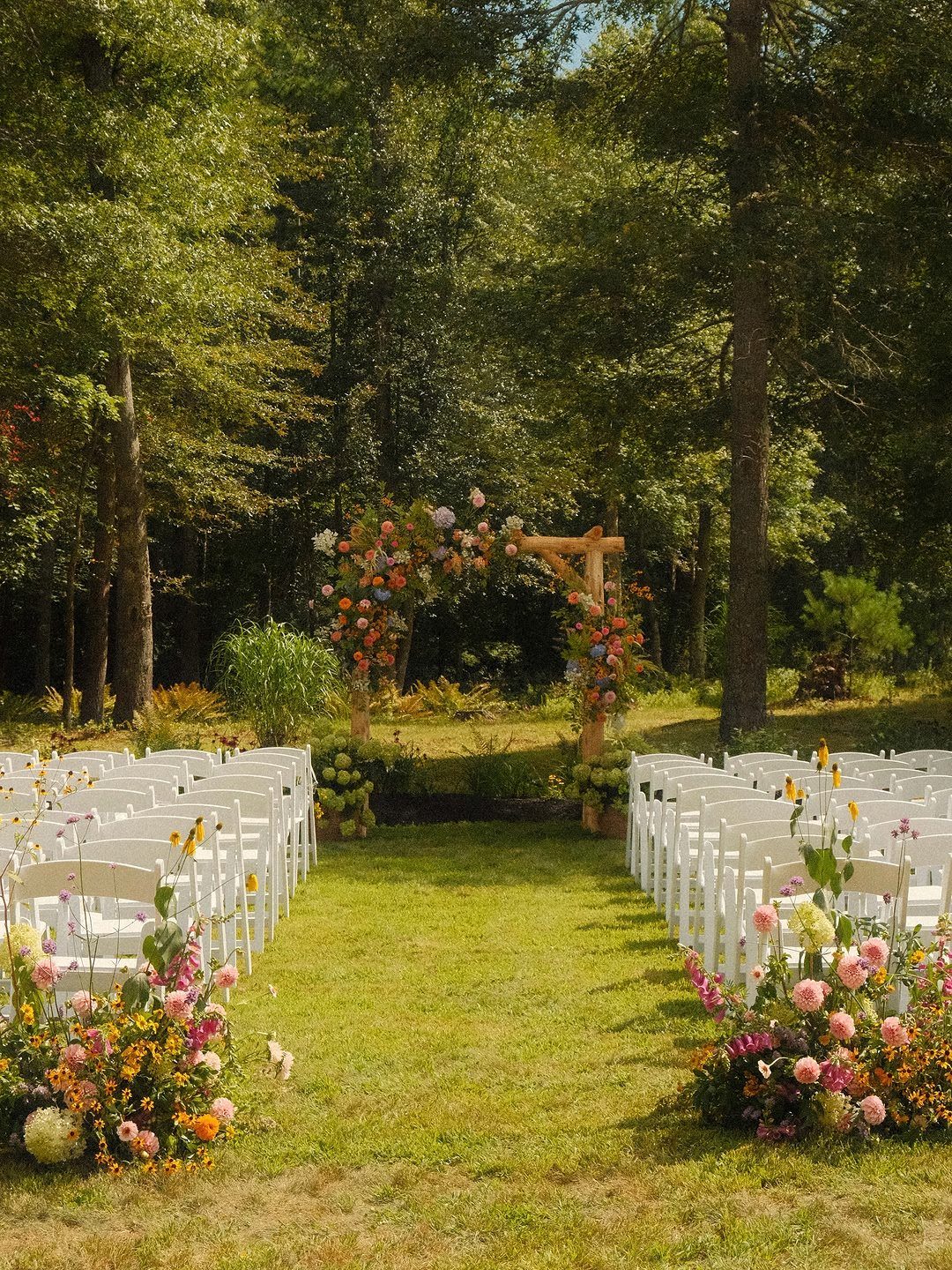 White wedding chairs arranged on grass facing a floral wooden arch in a forest.
