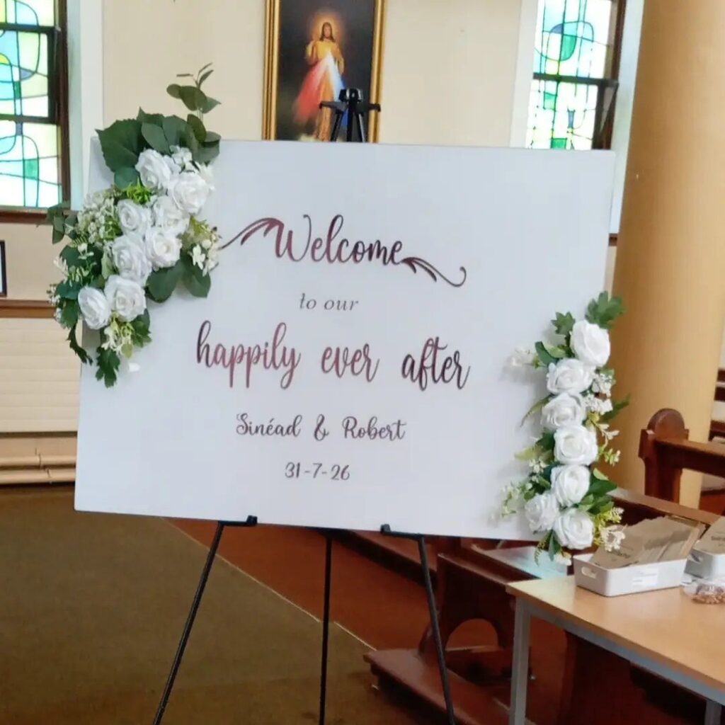 A white wedding welcome sign with floral decorations stands on an easel inside a church.