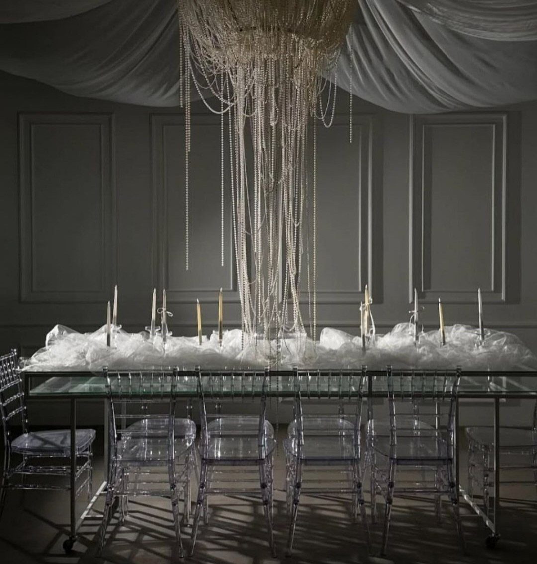 A long glass table decorated with white tulle and candles under a beaded chandelier.