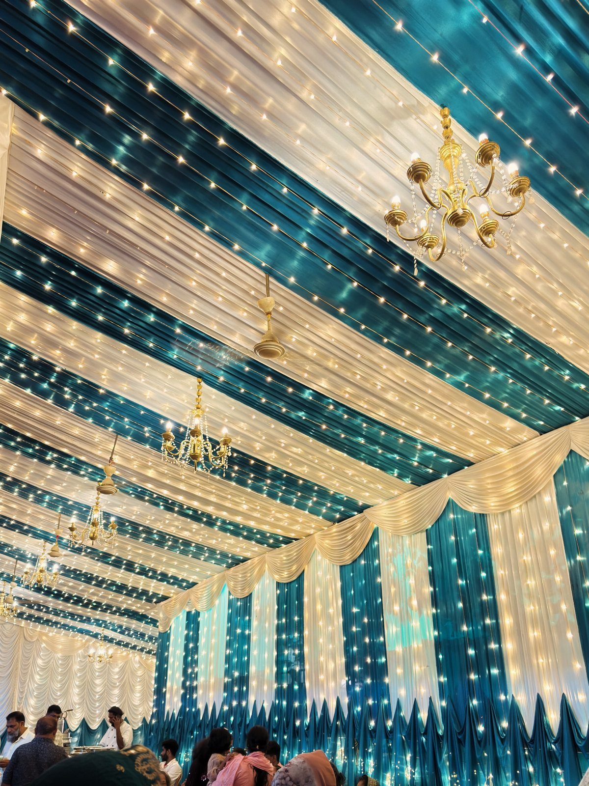 Teal and white striped fabric ceiling decorated with fairy lights and gold chandeliers in a tent.