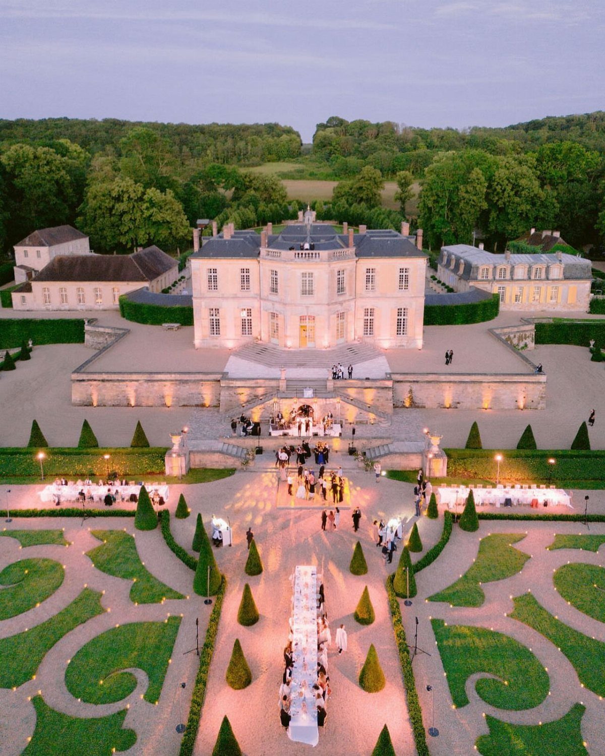 A large historic white manor house with formal gardens and outdoor wedding dining tables at twilight.