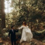A bride and groom walking through a sunlit forest with the bride stepping over a log.