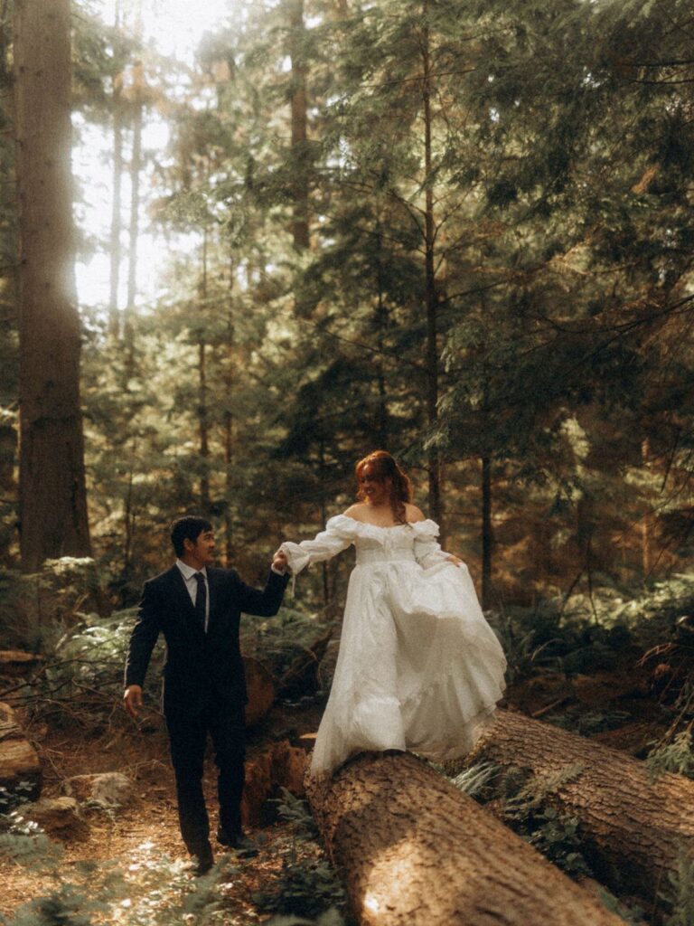 A bride and groom walking through a sunlit forest with the bride stepping over a log.