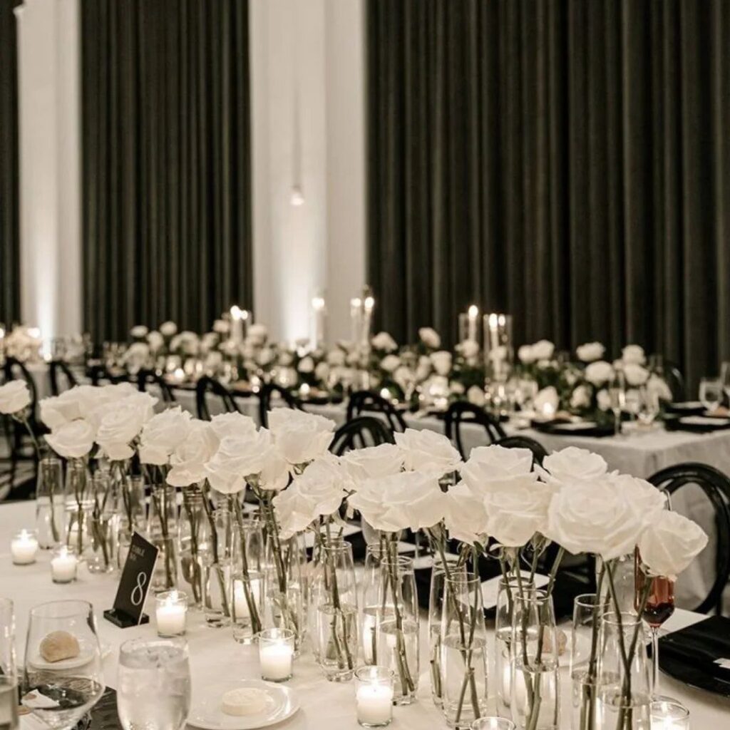 White roses in glass vases arranged on a long wedding reception table with candles.