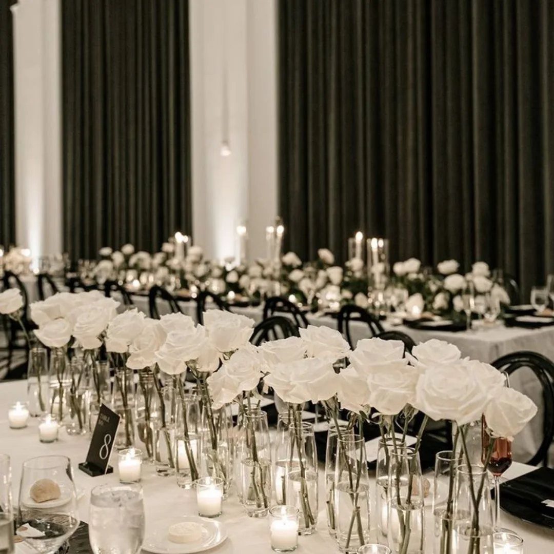 White roses in glass vases arranged on a long wedding reception table with candles.
