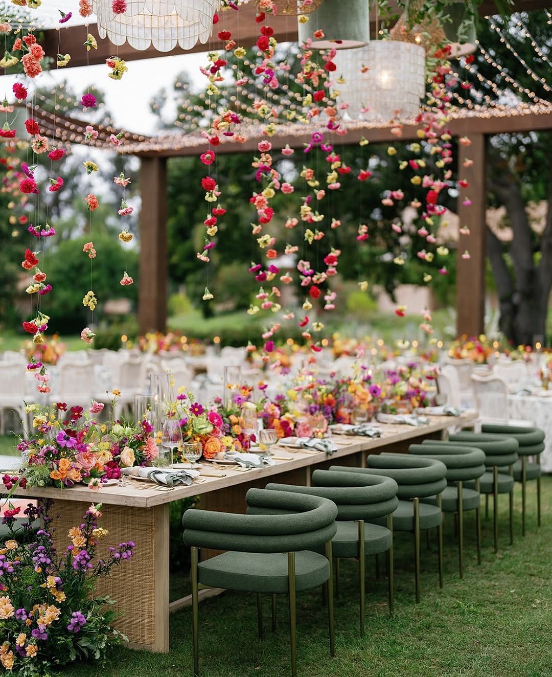 Long wooden dining table decorated with colorful flowers under a pergola in an outdoor garden setting.