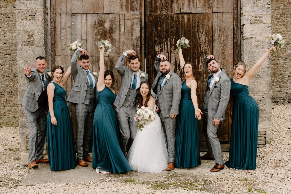 Wedding party posing in teal dresses and grey suits in front of wooden doors.