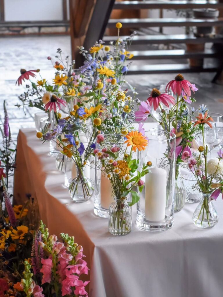 Colorful wildflowers in small glass vases arranged on a long wedding table with white candles.